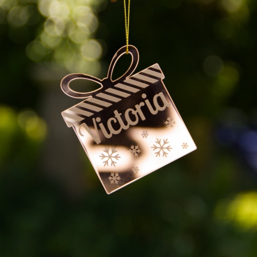 A gold mirror christmas ornament hanging from a gold string. It has the name Victoria etched into it, and the ornament is the shape of a present