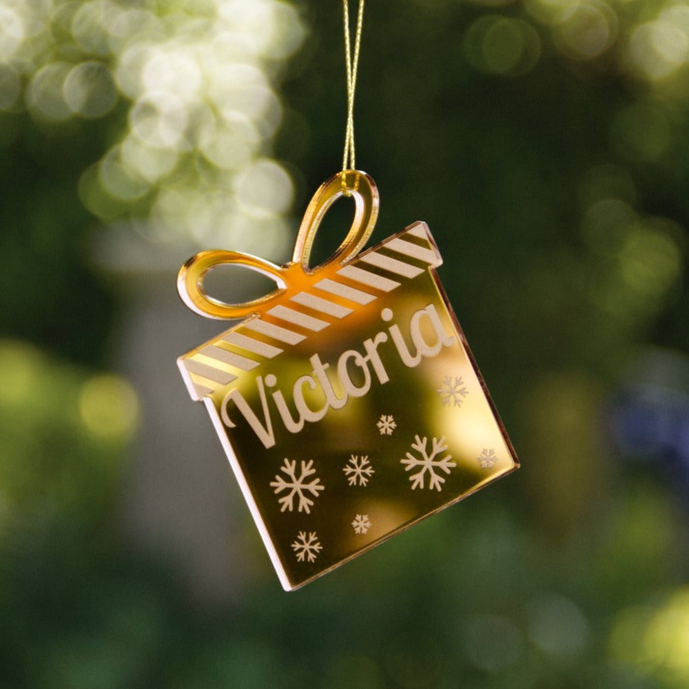 A mirror gold christmas ornament with the name Victoria etched. The ornament is the shape of a present.