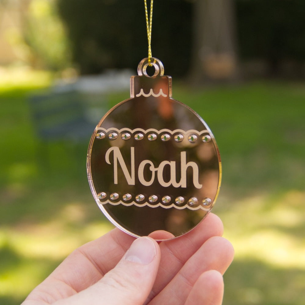 A mirror gold christmas ornament with the name Noah etched on to it. The ornament is the shape of a round bauble.