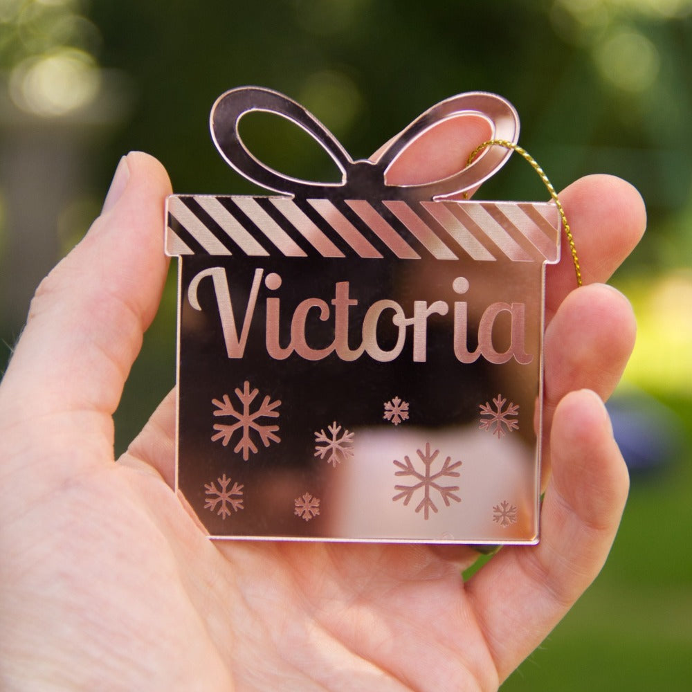 A person is holding a christmas ornament that is the shape of a present and is made from mirror gold acrylic. It is personalised with the name Victoria etched on to it.