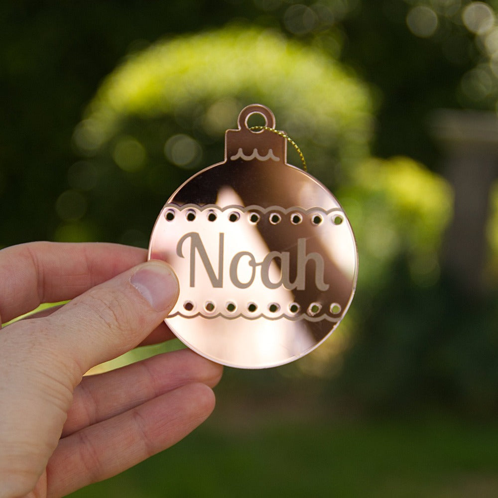 A person is holding a gold mirror christmas ornament that is personalised with the name Noah