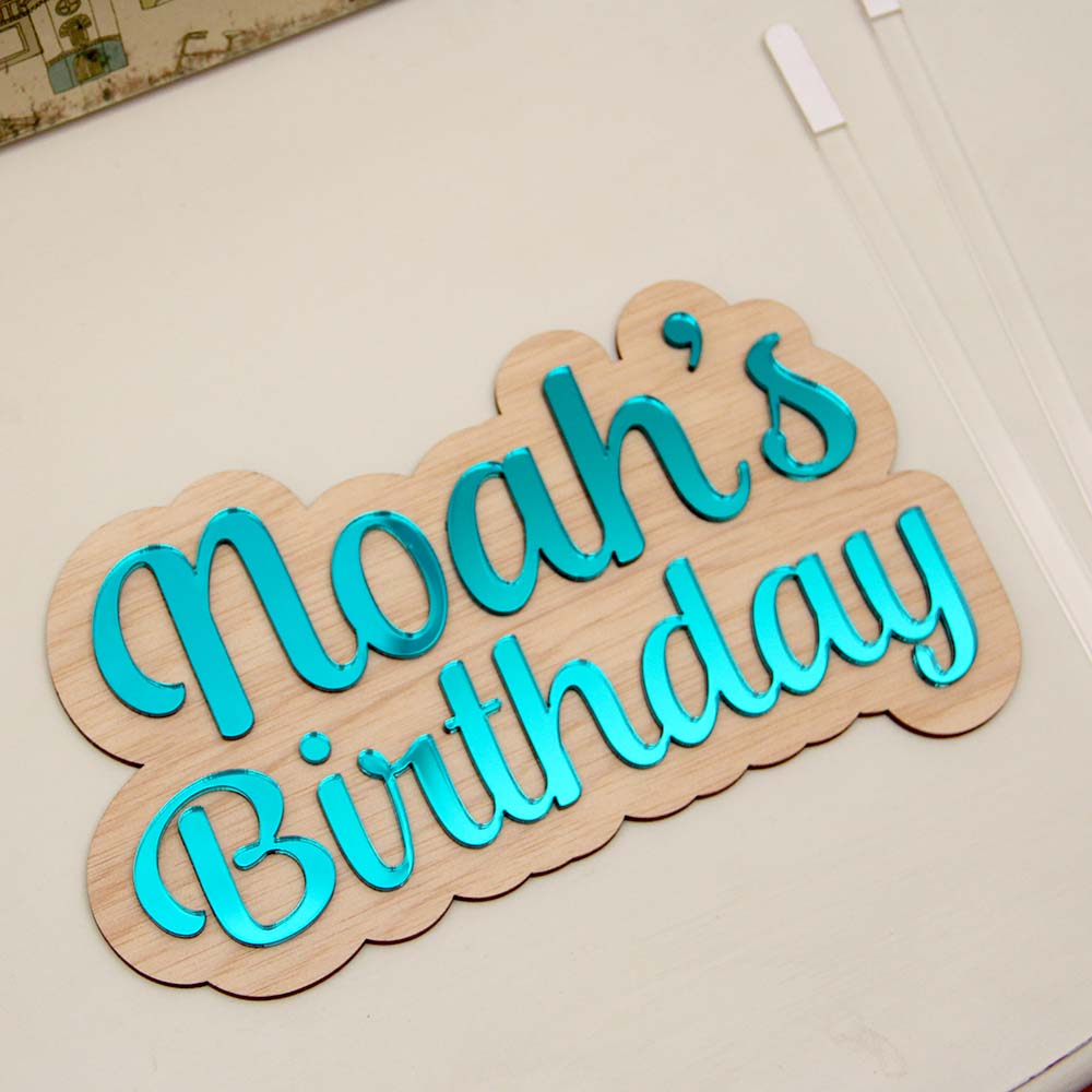Wood plaque with "noah's birthday" written in blue mirror acrylic. Clear sticks will be stuck on to make a cake topper