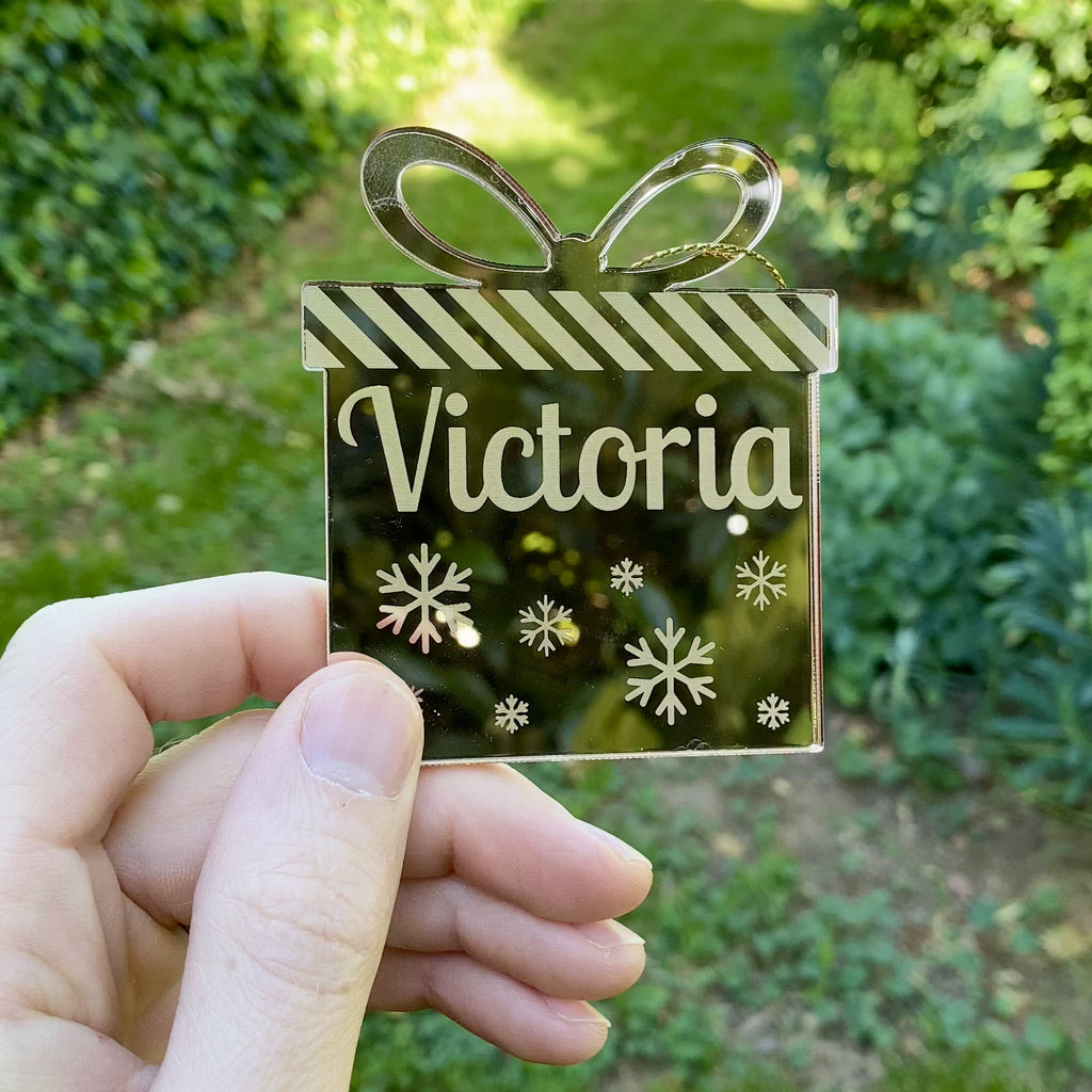 A video showing a person holding a mirror gold Christmas ornament that is personalised with the name Victoria. The ornament is being moved around to reflect the garden.
