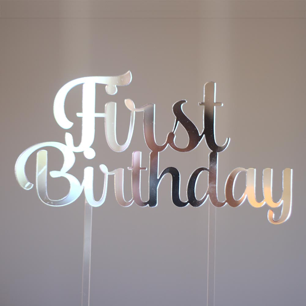 A silver mirror cake topper that says First Birthday. It has two clear sticks that go into the cake