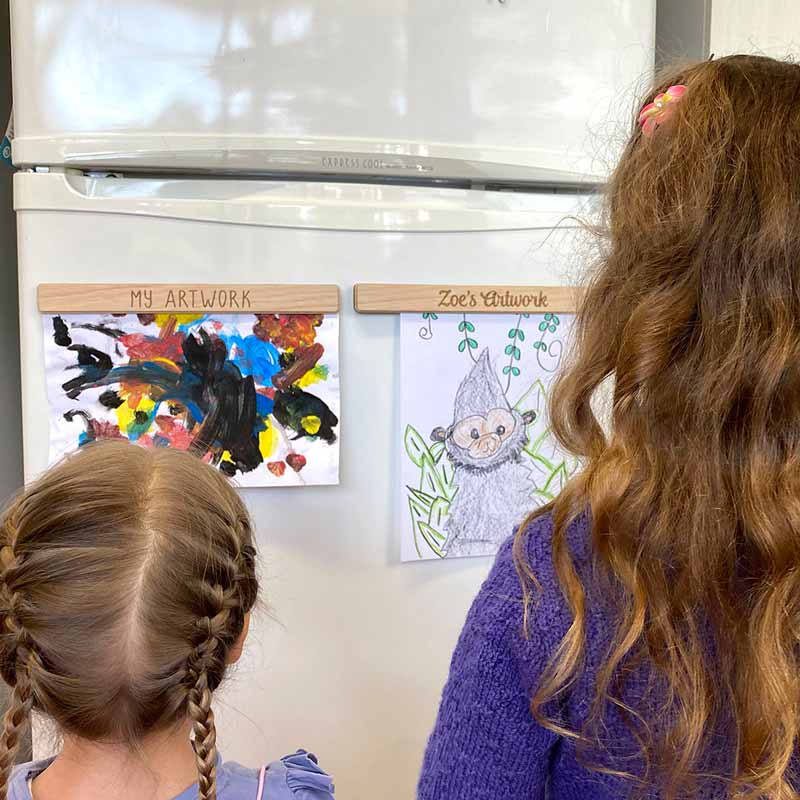 two girls facing away from the camera looking at their artwork hanging on the fridge
